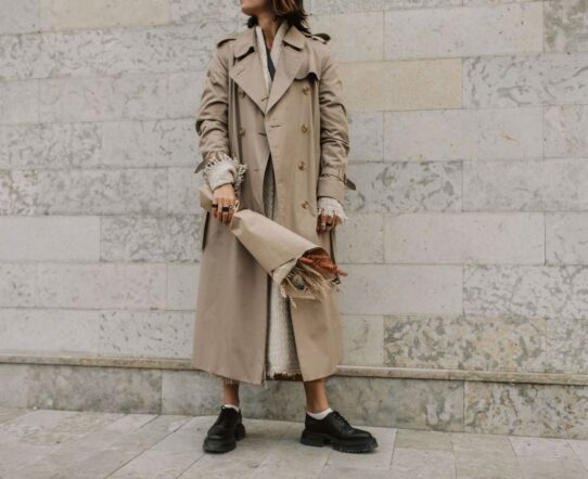 Person i beige trenchcoat, stickad tröja och kraftiga svarta skor