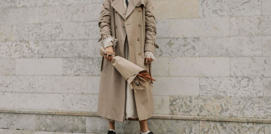 Person i beige trenchcoat, stickad tröja och kraftiga svarta skor