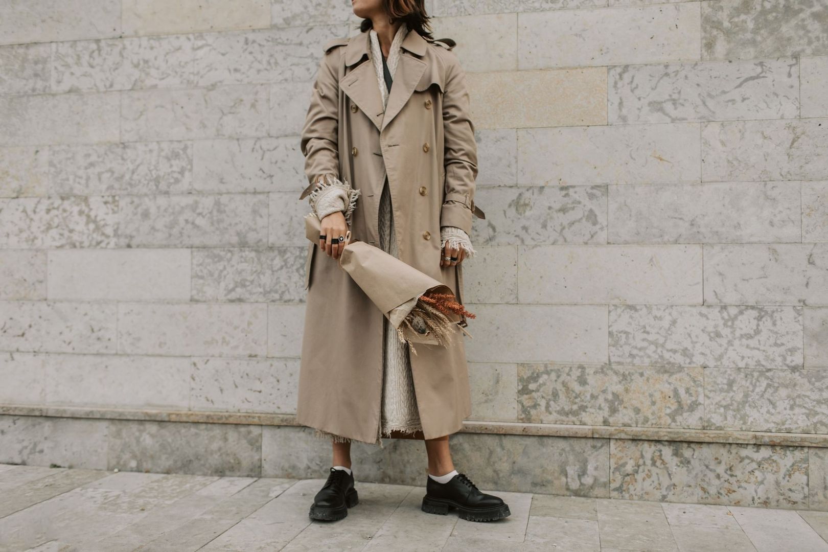 Person i beige trenchcoat, stickad tröja och kraftiga svarta skor